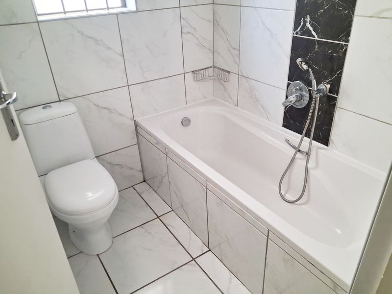 A clean, modern bathroom featuring white marble-look wall and floor tiles, a built-in bathtub with a shower mixer, and a standard toilet.