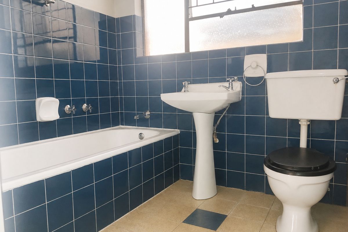 A functional bathroom featuring a pedestal basin, bathtub, and toilet, finished with dark blue wall tiling and a window with security bars.
