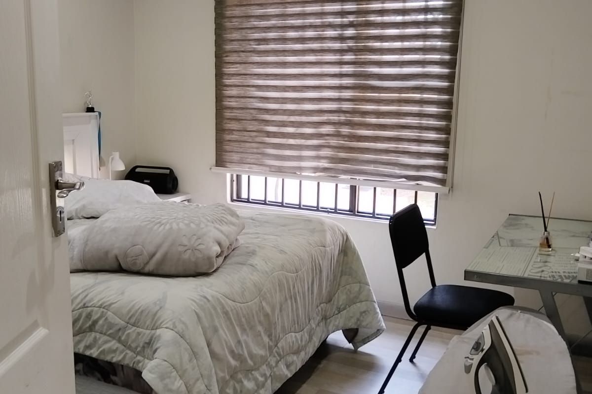 A functional bedroom featuring light-toned laminate flooring, decorative ceiling cornicing, and a window fitted with modern roller blinds and security bars.