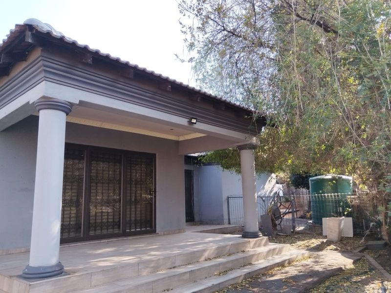 A covered patio area featuring tiled flooring, structural columns, and sliding glass doors with security gates, overlooking a garden space with a water tank.