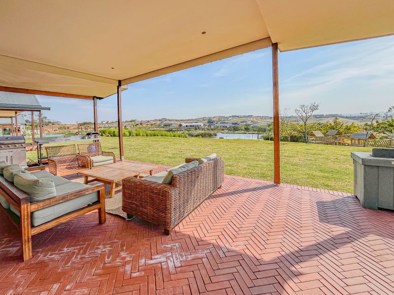 A spacious covered patio featuring herringbone brick paving, timber support posts, and a built-in braai, overlooking a large lawn and scenic landscape.