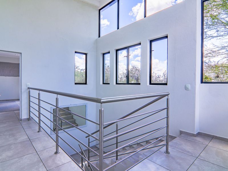 A bright, double-volume landing area featuring modern stainless steel balustrades, large windows providing ample natural light, and neutral tiled flooring.