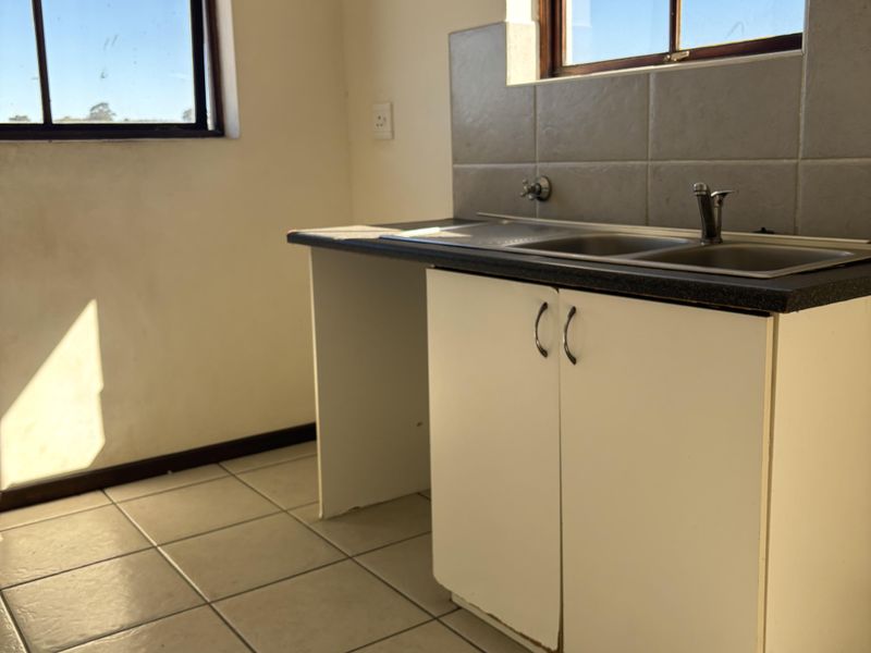 A compact, empty kitchen featuring a stainless steel sink, tiled splashback, and basic base cabinetry with tiled flooring.