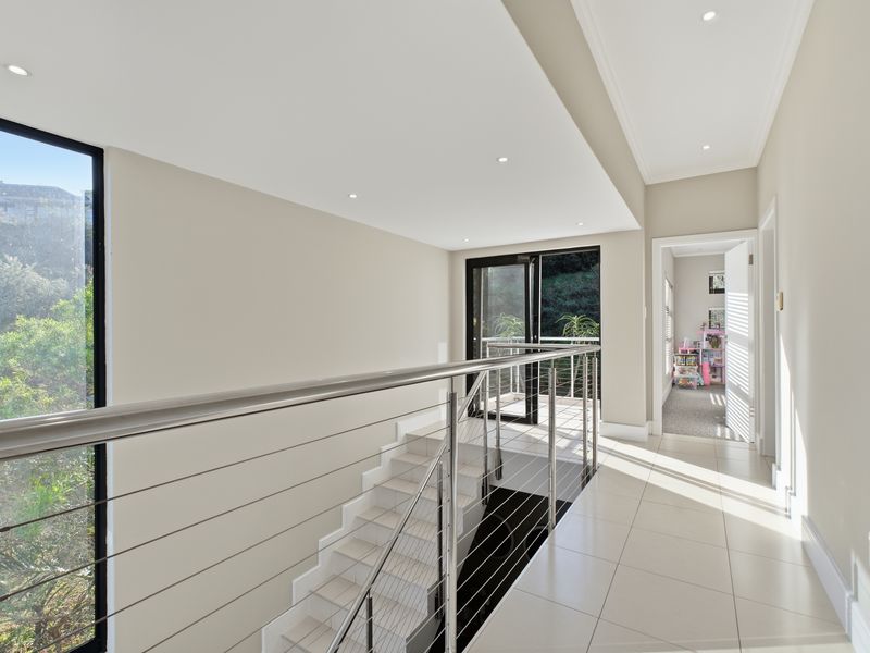 Bright and modern upper-level hallway featuring tiled flooring, a sleek stainless steel cable balustrade, and large windows providing natural light and garden views.
