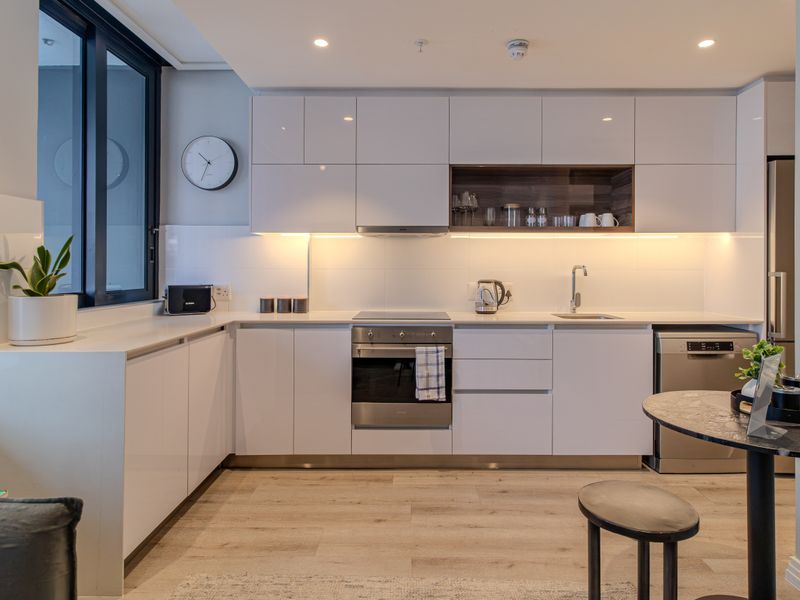 Modern kitchen featuring sleek white cabinetry, integrated appliances, an electric hob, and under-cabinet lighting, finished with light wood-look flooring.
