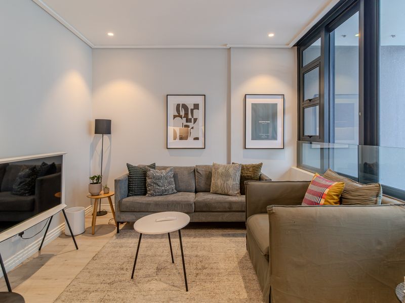 A modern, well-lit living room featuring large windows with a glass balustrade, recessed lighting, and light-toned laminate flooring.