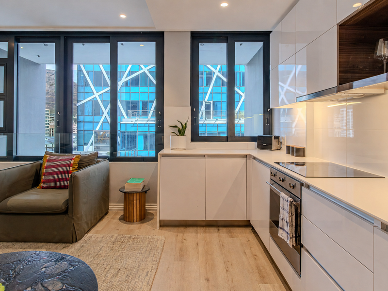 Modern open-plan kitchen and living area featuring sleek white cabinetry, integrated appliances, stone countertops, and large windows providing ample natural light.