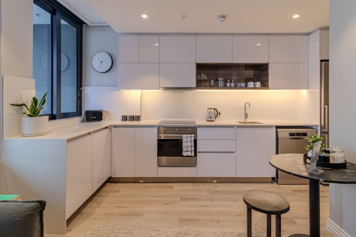 Modern kitchen featuring sleek white cabinetry, integrated appliances, under-cabinet lighting, and light wood-look flooring in a clean, contemporary apartment setting.