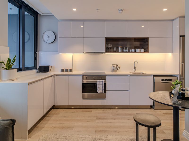 Modern kitchen featuring sleek white cabinetry, integrated appliances, under-cabinet lighting, and light wood-look flooring in a clean, contemporary apartment setting.