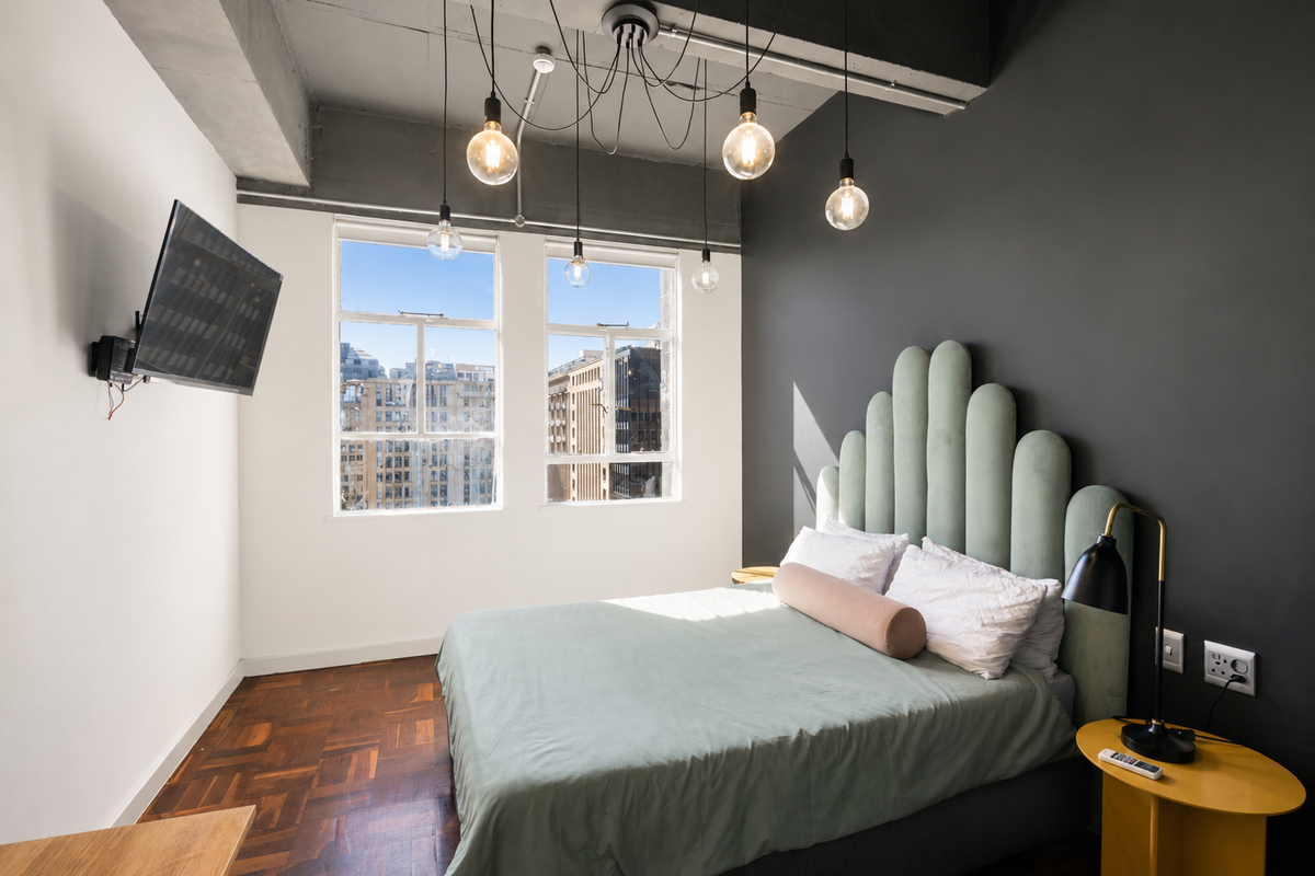 Modern bedroom featuring parquet flooring, an exposed concrete ceiling with industrial pendant lighting, and large windows offering city views.