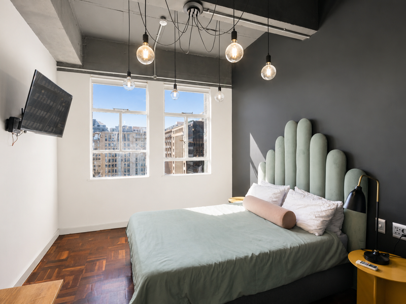 Modern bedroom featuring parquet flooring, an exposed concrete ceiling with industrial pendant lighting, and large windows offering city views.