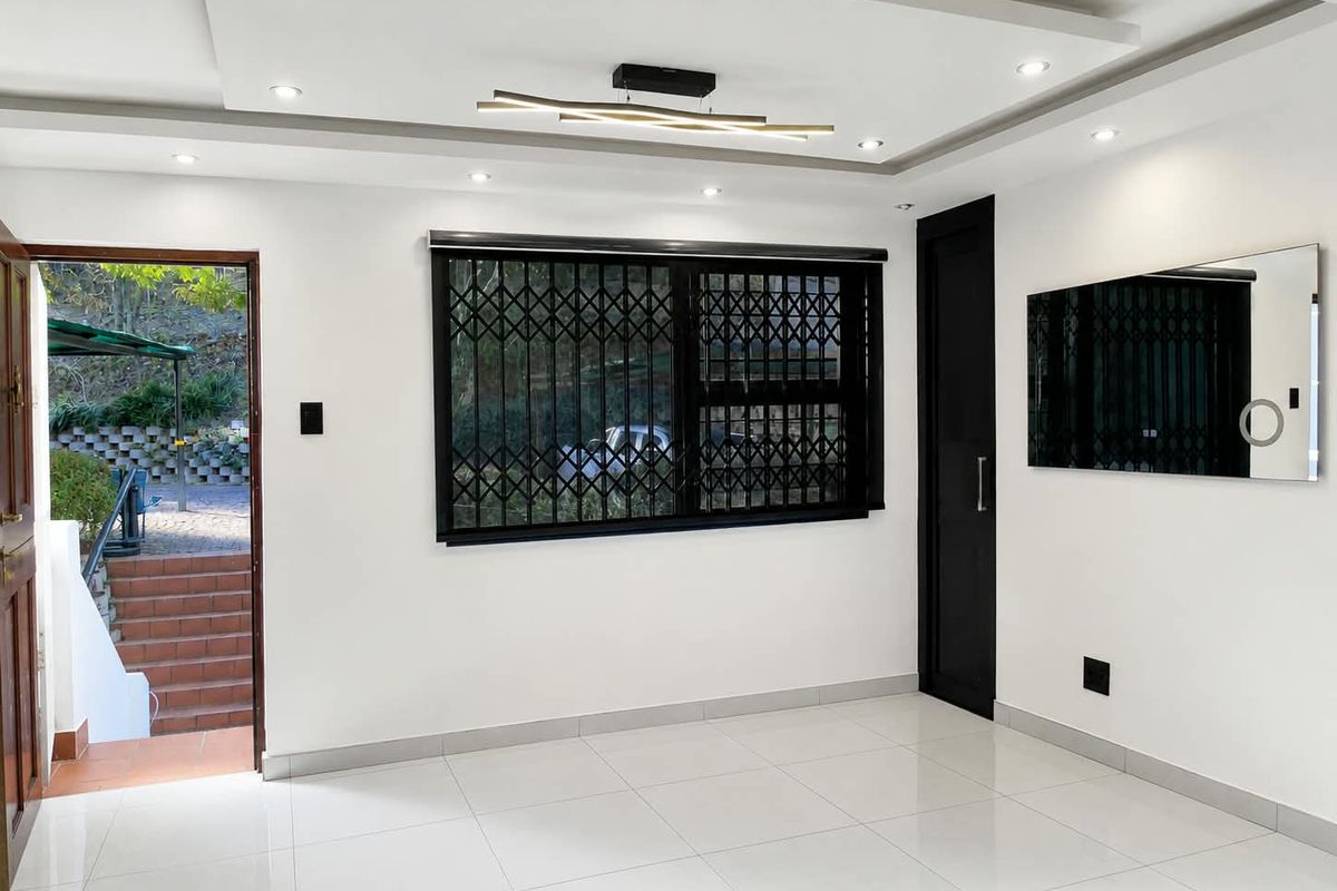 A bright, empty living room featuring modern recessed ceiling lighting, white tiled floors, and a secure window with a fitted security gate.