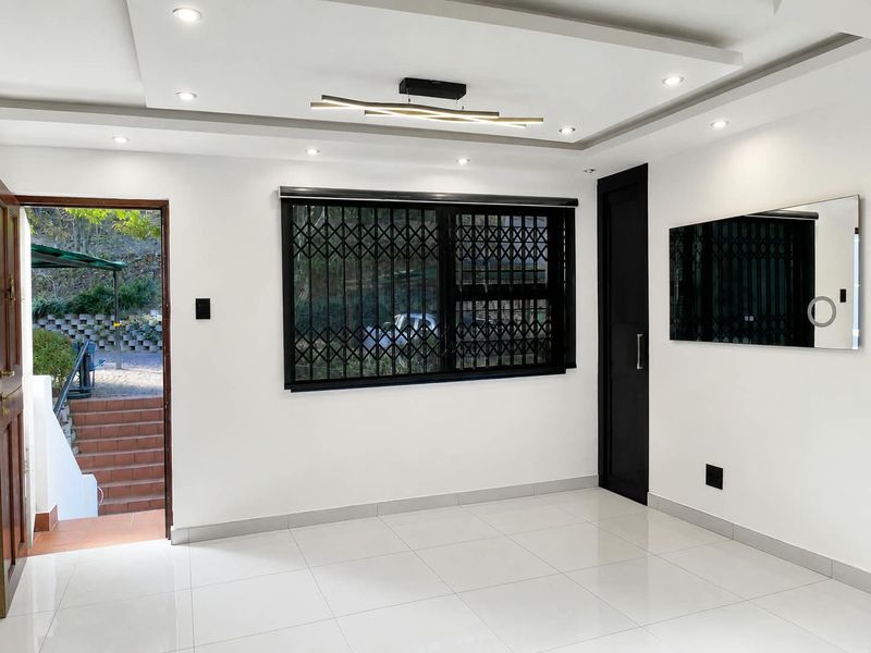 A bright, empty living room featuring modern recessed ceiling lighting, white tiled floors, and a secure window with a fitted security gate.