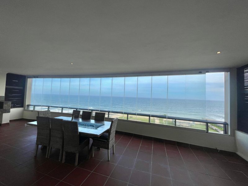 Spacious covered balcony featuring tiled flooring, a built-in braai, and a frameless glass enclosure offering panoramic ocean views.
