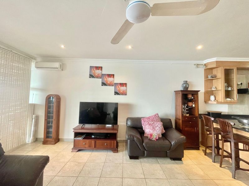 Open-plan living area featuring tiled flooring, a ceiling fan, and a split-system air conditioner, seamlessly connecting to a kitchen with a breakfast bar.