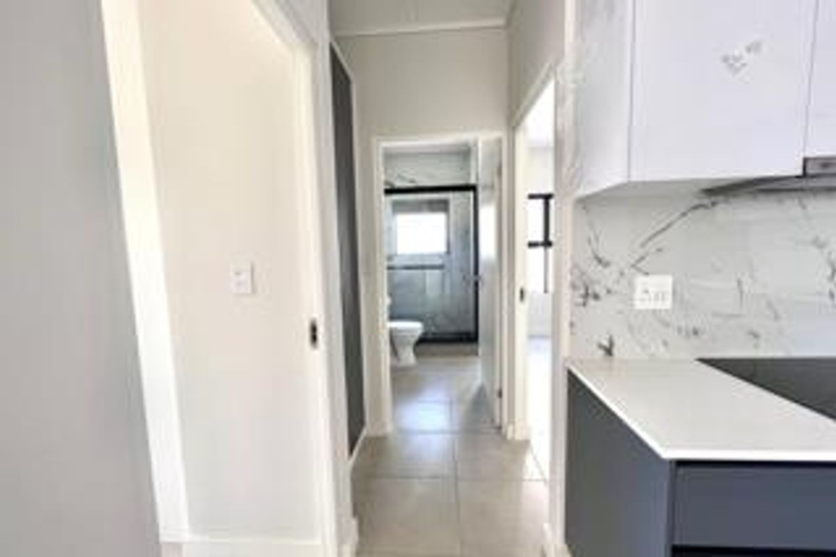A bright hallway connecting to a modern kitchen with stone countertops and marble-look splashbacks, leading towards a bathroom and bedroom area.