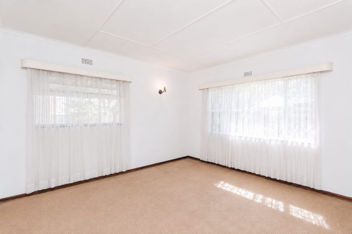 A bright, empty living room featuring neutral carpet flooring, classic ceiling tiles, and dual windows with fitted curtain pelmets.
