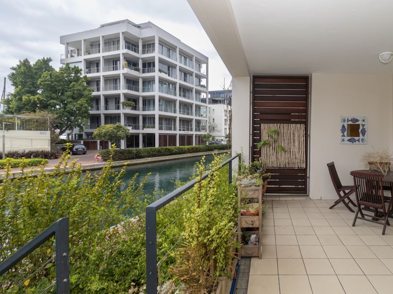 A covered balcony featuring tiled flooring, a metal balustrade, and a timber privacy screen, offering views over the canal and surrounding apartment buildings.