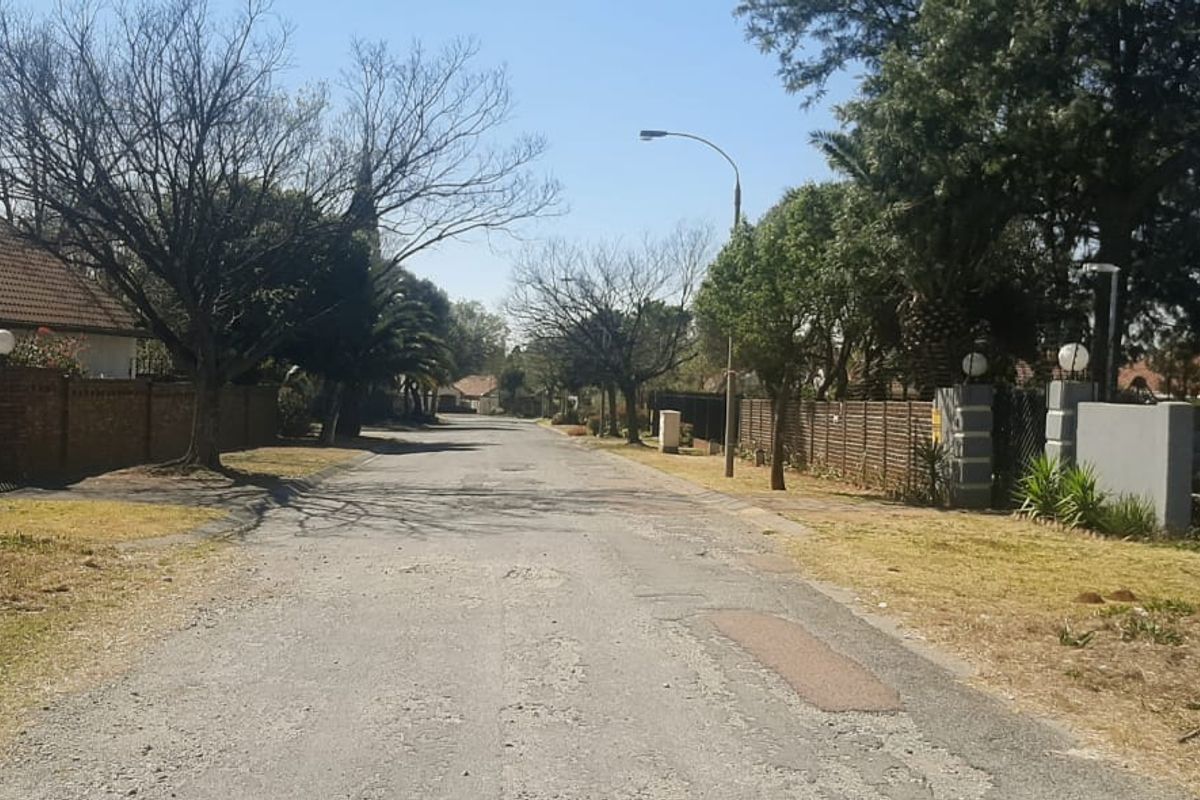 A view of the residential street showing the asphalt road, surrounding boundary walls, and established trees in a suburban neighborhood setting.