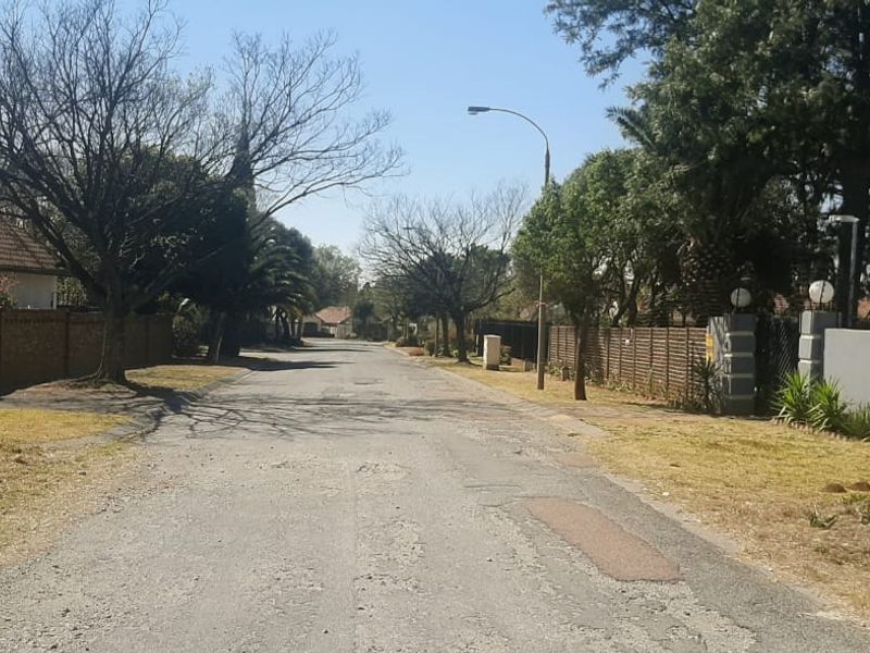 A view of the residential street showing the asphalt road, surrounding boundary walls, and established trees in a suburban neighborhood setting.