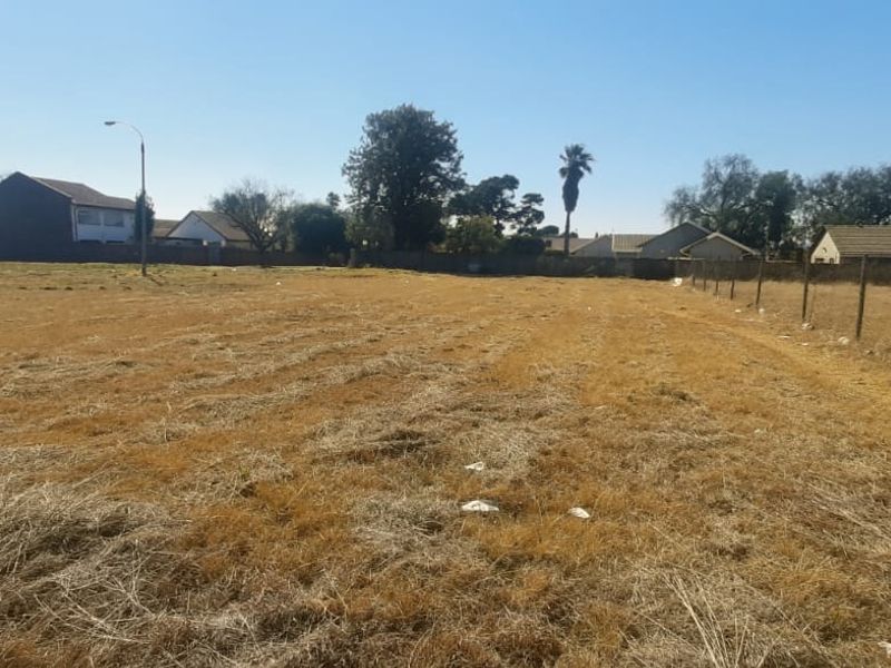 A vacant residential plot of land featuring dry grass, a wire perimeter fence, and surrounding neighborhood views.