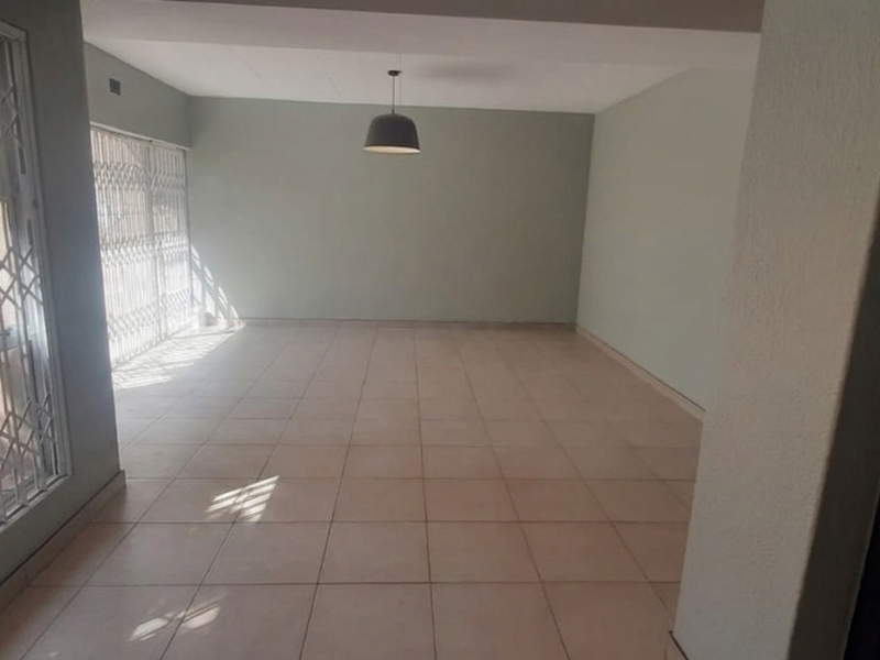 A spacious, empty living area featuring neutral tiled flooring, security gates on the windows, and a central pendant light fixture.