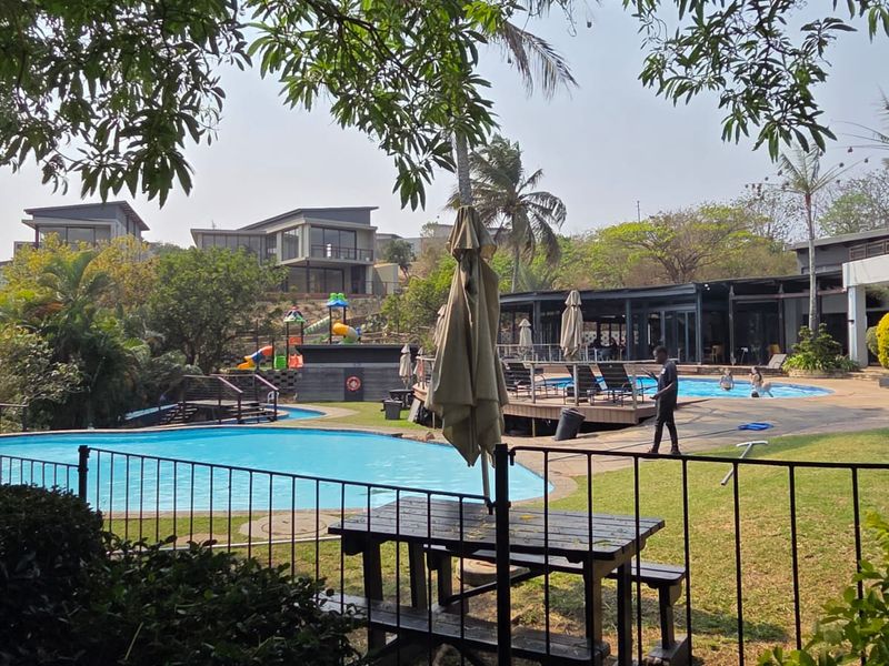 A communal outdoor pool area featuring a swimming pool, wooden decking, and a children's play area within a landscaped residential estate setting.
