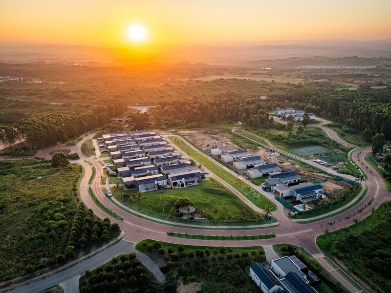 Aerial view of a modern residential estate development featuring paved roads, landscaped grounds, and a tennis court at sunset.