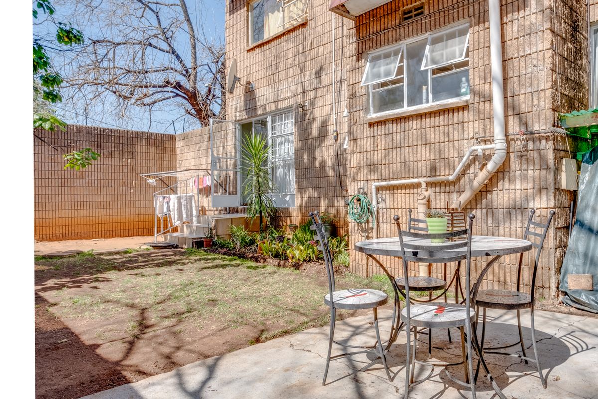 A rear exterior view of the apartment featuring a private garden area, concrete patio, and brick facade with casement windows.
