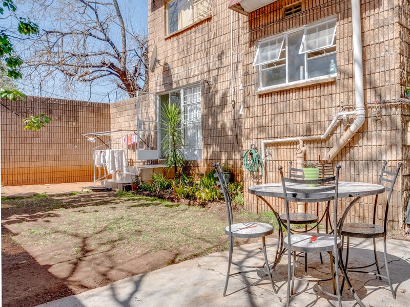A rear exterior view of the apartment featuring a private garden area, concrete patio, and brick facade with casement windows.