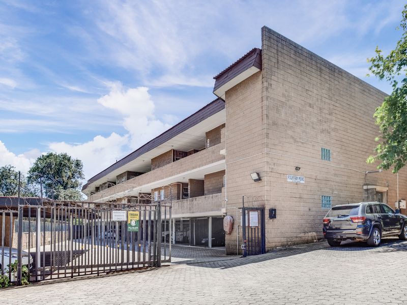 Exterior view of the Fountain Mews apartment complex featuring a brick facade, secure gated entrance, and paved driveway access.