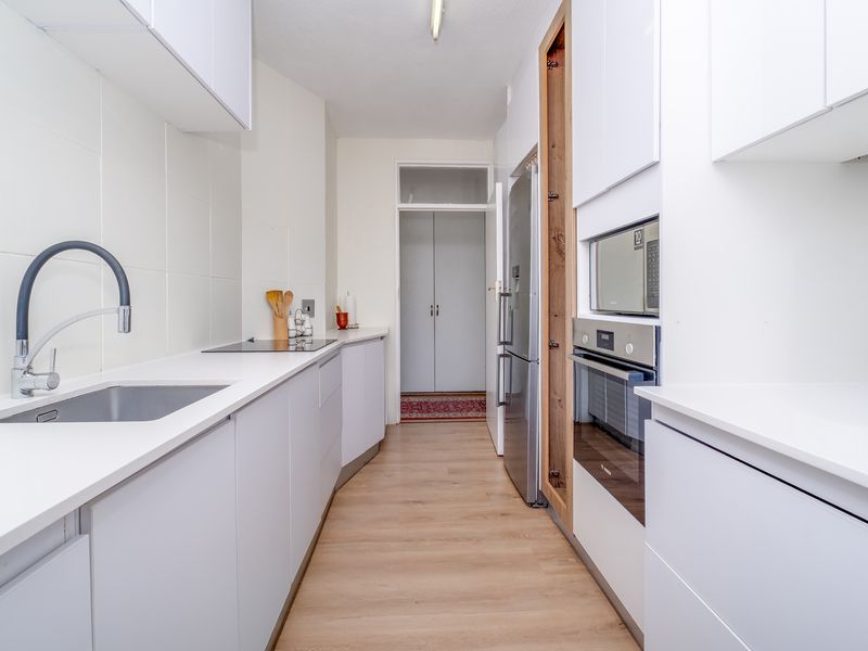 A modern, galley-style kitchen featuring white cabinetry, integrated appliances including an oven and microwave, and a sleek stainless steel sink with a contemporary tap.