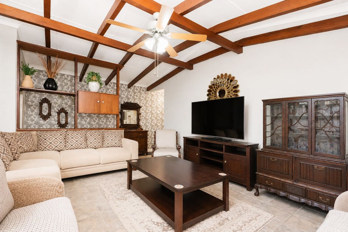 A living room featuring exposed timber ceiling beams, a ceiling fan, tiled flooring, and a built-in shelving unit with decorative wallpaper.