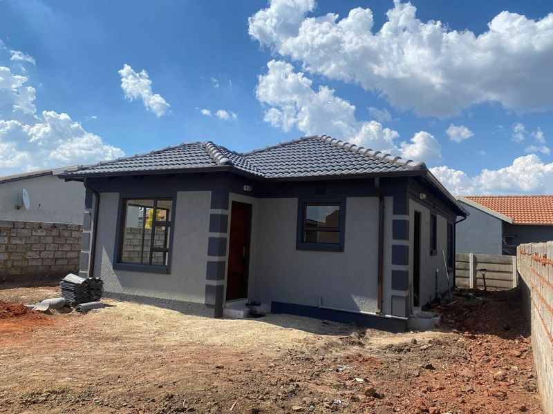 A newly constructed single-story house featuring a modern grey facade, tiled roof, and aluminium window frames, set on an unfinished plot.