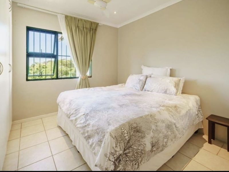 A functional bedroom featuring tiled flooring, built-in cupboards, a ceiling fan, and a window fitted with security bars.