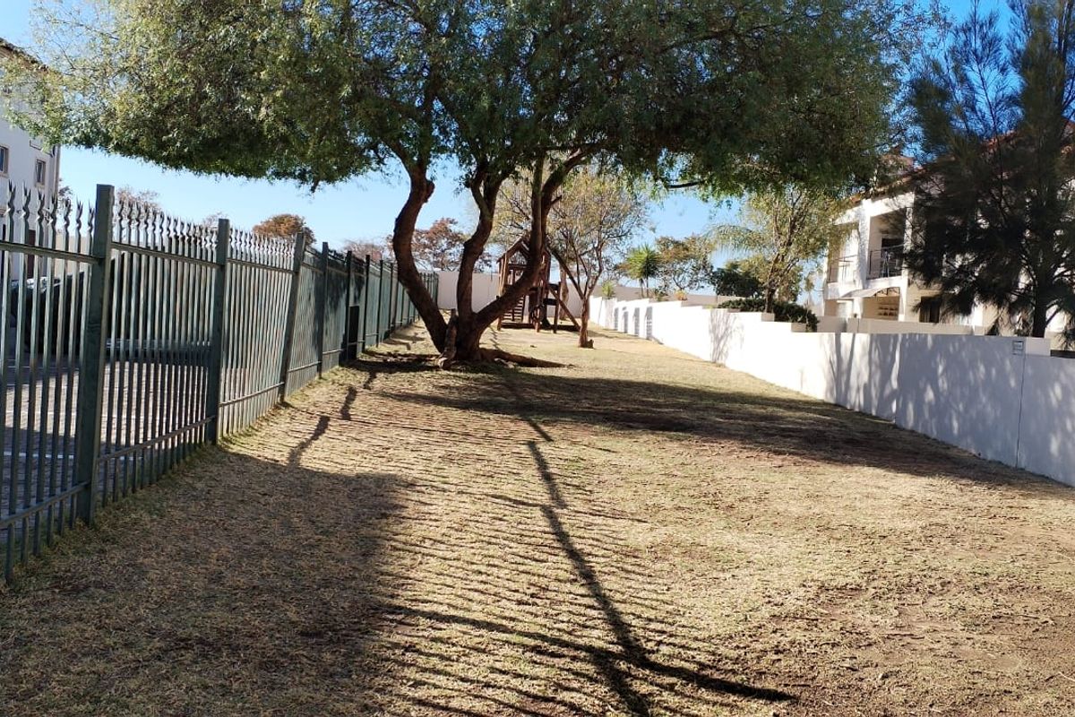 A view of the apartment complex grounds featuring a large lawn area, mature trees, and secure perimeter fencing.