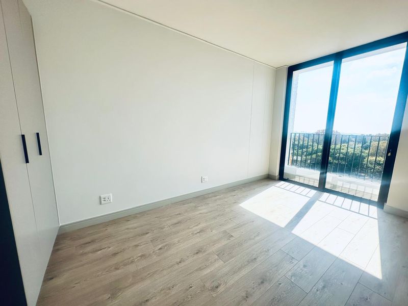 A bright, empty bedroom featuring modern laminate flooring, a built-in wardrobe, and large sliding glass doors leading to a balcony with scenic views.