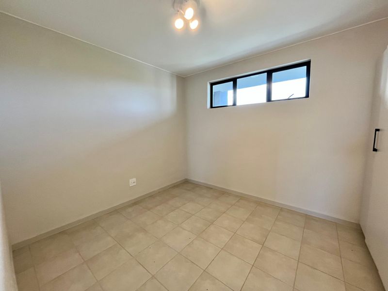 A clean, empty bedroom featuring neutral tiled flooring, a built-in cupboard, and a high-level window providing natural light.