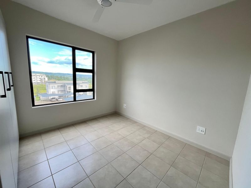 A bright, empty bedroom featuring neutral tiled flooring, built-in cupboards, a ceiling fan, and a large window with views of the surrounding area.