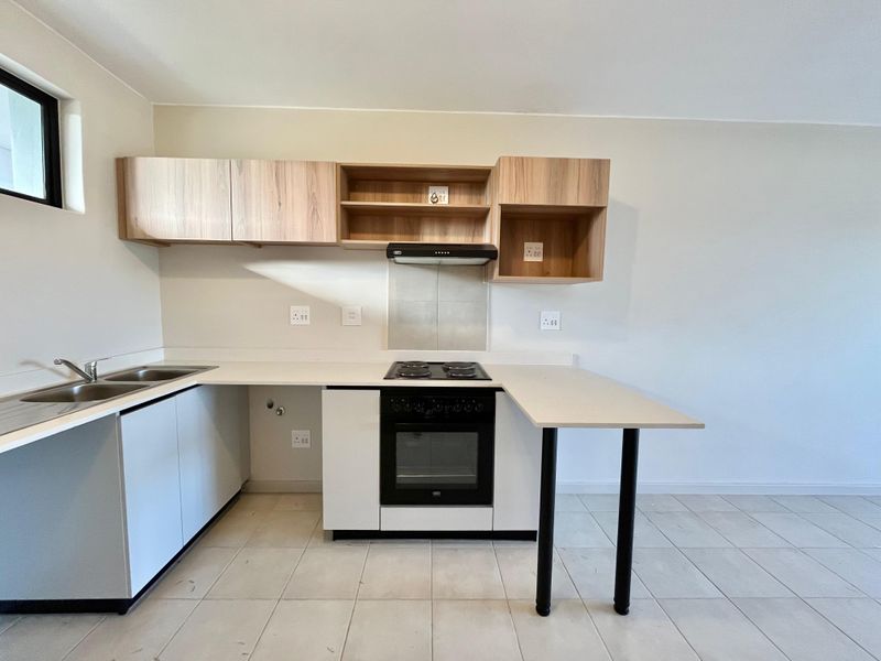 A clean, modern kitchen featuring light wood-tone cabinetry, white stone countertops, a built-in oven, hob, and extractor fan with neutral tiled flooring.
