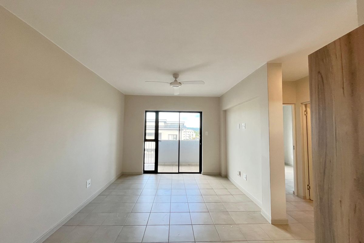 A bright, empty living room featuring neutral ceramic floor tiles, a ceiling fan, and a sliding glass door leading to an outdoor balcony area.