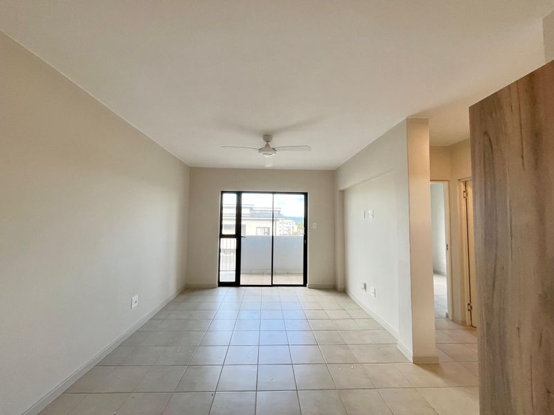 A bright, empty living room featuring neutral ceramic floor tiles, a ceiling fan, and a sliding glass door leading to an outdoor balcony area.