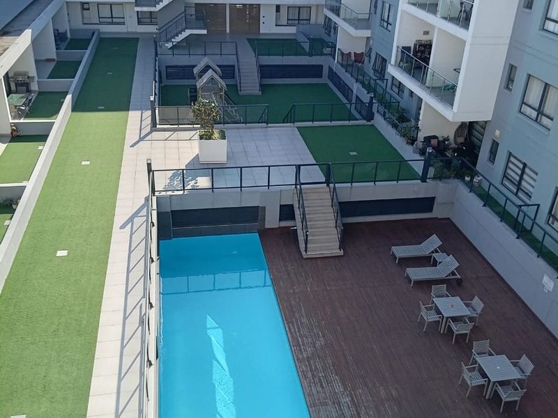 An elevated view of a modern apartment complex courtyard featuring a swimming pool, timber deck, artificial lawn areas, and secure glass balustrades.