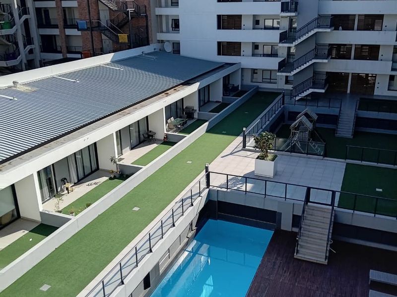 Aerial view of an apartment complex courtyard featuring a swimming pool, wooden decking, artificial grass areas, and multi-level building access.