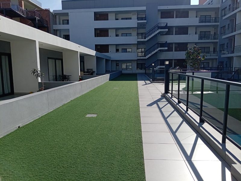 A well-maintained communal outdoor area featuring artificial turf, tiled pathways, and modern glass balustrades within an apartment complex.