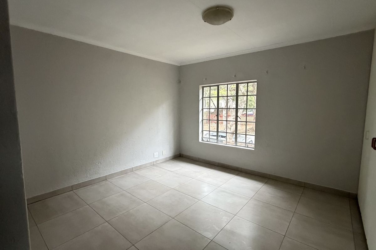 A bright, empty bedroom featuring neutral tiled flooring, built-in cupboards, and a window fitted with security burglar bars.