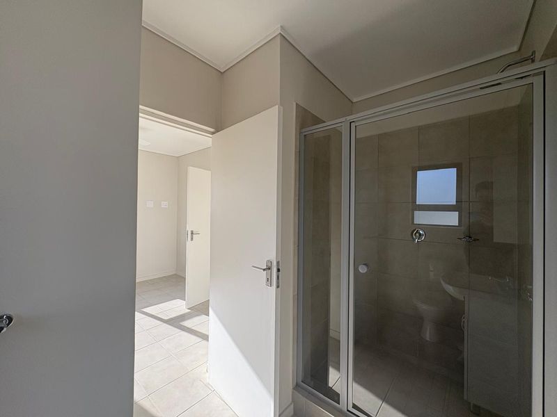 A clean, modern bathroom featuring a glass-enclosed shower, tiled finishes, and neutral tones, adjacent to a bright, empty living space.