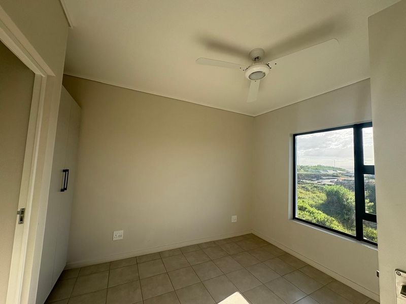 A bright, empty bedroom featuring neutral tiled flooring, a built-in cupboard, a ceiling fan, and a large window with scenic views.