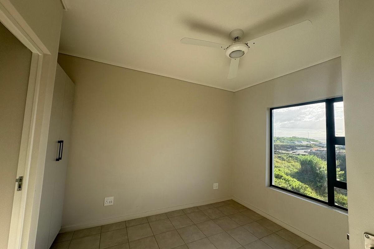 A bright, empty bedroom featuring neutral tiled flooring, a built-in cupboard, a ceiling fan, and a large window with scenic views.