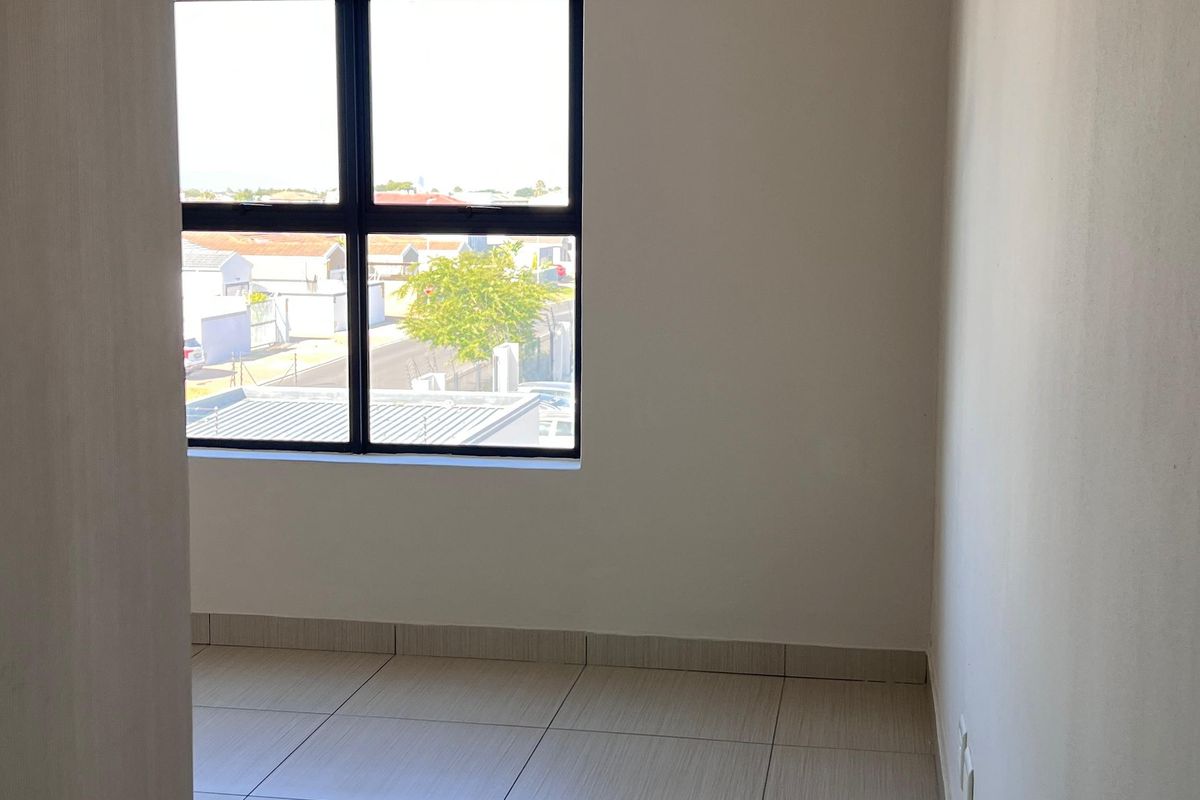 A bright, empty bedroom featuring neutral tiled flooring and a modern aluminium-framed window with a view of the surrounding neighbourhood.